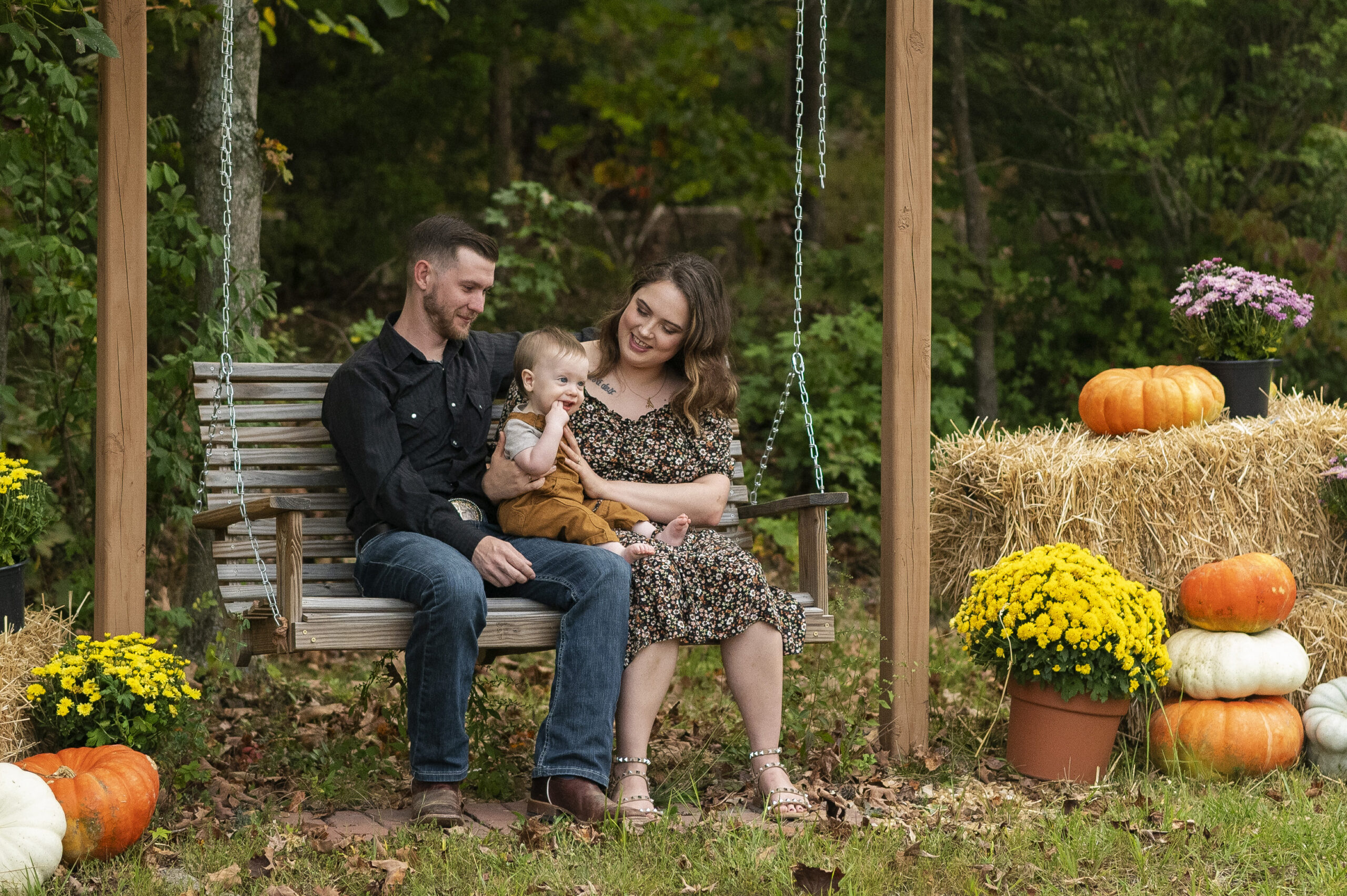 family of three sits on wooden swing with fall decor around them